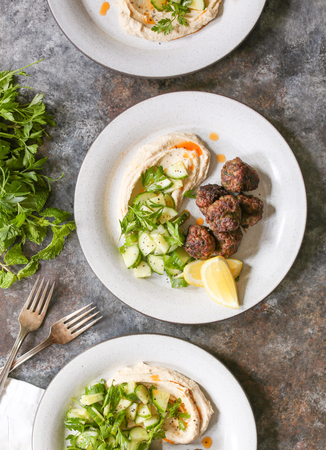 Overhead view of a plated serving of lamb kofta with hummus, cucumbers, and lemon wedges.