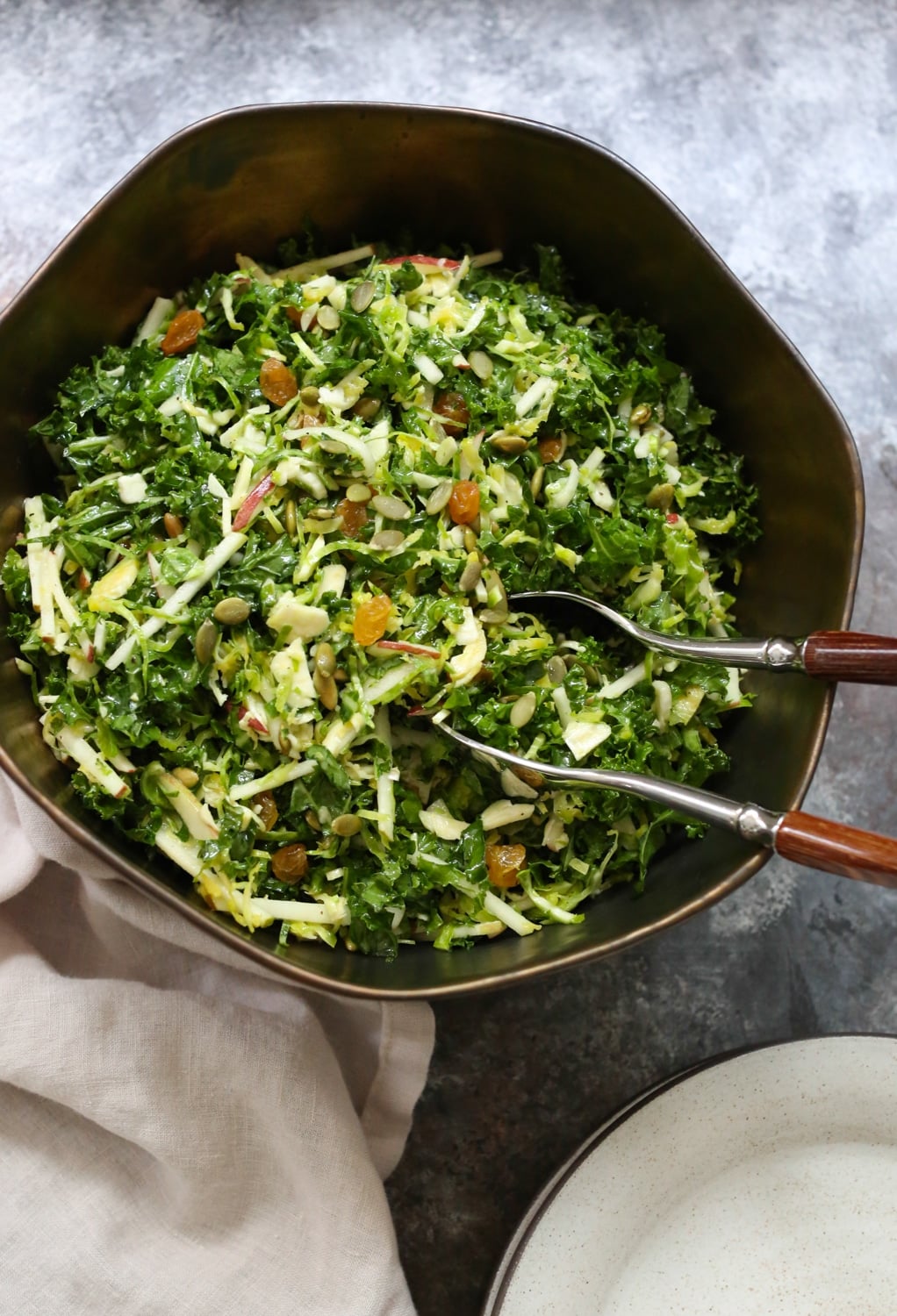 Overhead closeup of Kale, Brussels Sprout, and Apple Salad in a bowl with serving utensils in the bowl and side plates in the bottom right corner.